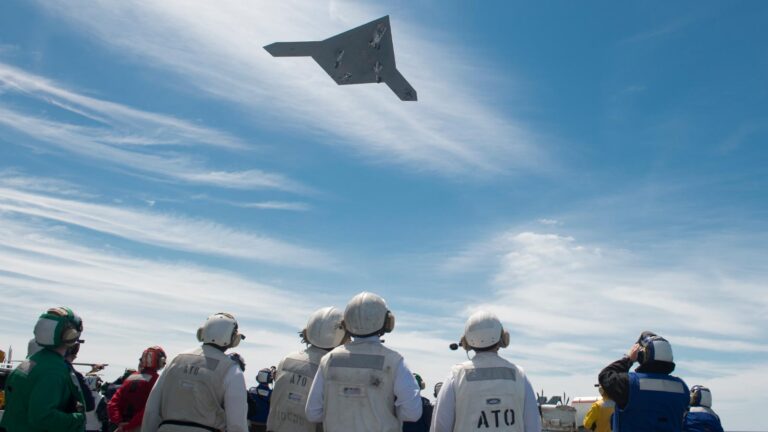 A group of people wearing helmets and protective gear watch a flying aircraft in a clear blue sky with scattered clouds.