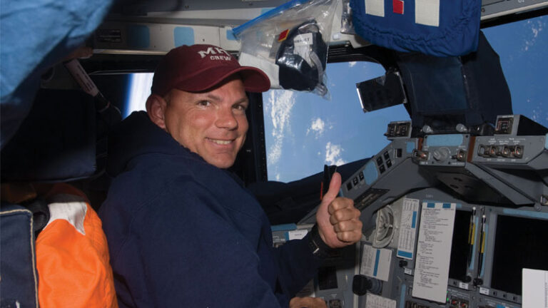 Astronaut inside a spacecraft giving a thumbs up, surrounded by control panels and a window showing the earth's atmosphere.