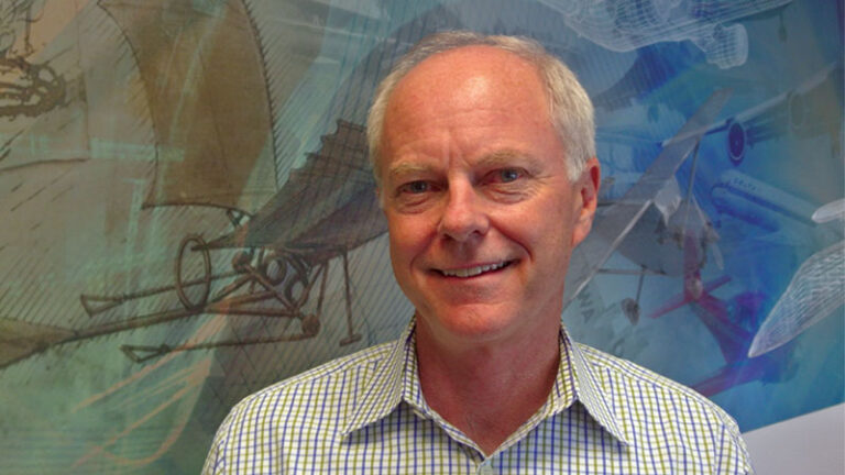 A middle-aged man with a smile, wearing a checkered shirt, standing in front of a wall with assorted aviation images.