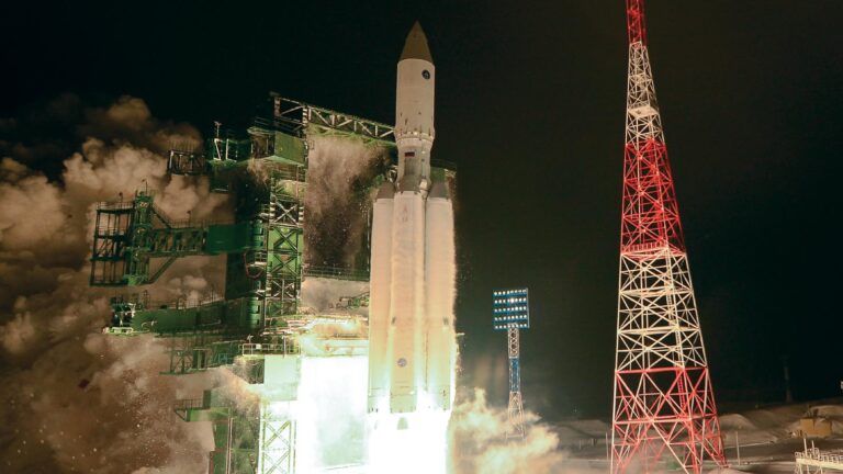 A rocket launching into the night sky from a platform, with large amounts of smoke and steam visible, and a triangular red and white structure to the right.