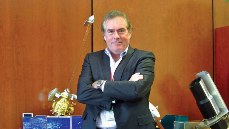 A man with grey hair, wearing a white shirt and red tie, stands with crossed arms in an office surrounded by satellite models and space-related photos.