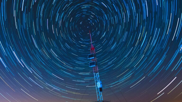 Long-exposure photograph of a telecommunications tower with circular star trails in the night sky, indicating the rotation of the Earth. Light streaks surround the tower, creating a vortex effect.