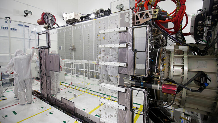 A technician in a clean suit inspects a large satellite instrument in a high-tech laboratory.