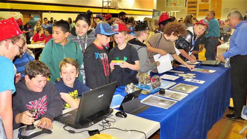 A group of children engaging with laptops and other materials at a busy school event in a gymnasium.