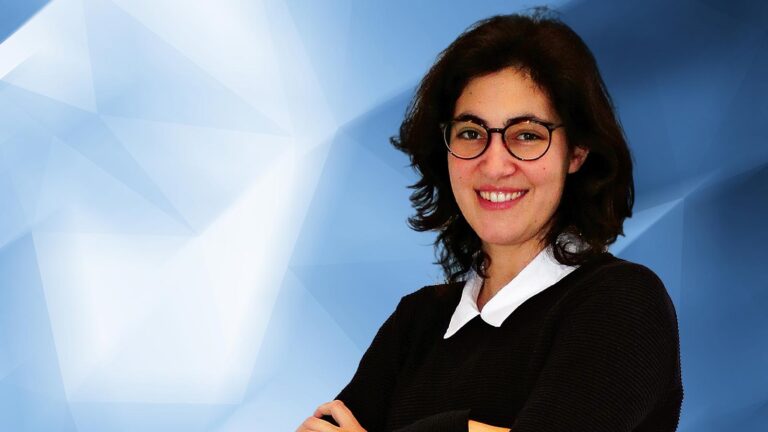 Person with short dark hair, wearing glasses and a black top with a white collar, smiling at the camera against a geometric blue and white background.