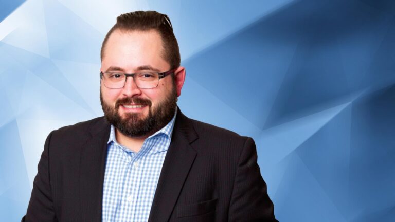 A man with a beard, wearing glasses, a black suit jacket, and a blue checkered shirt, stands against a blue geometric background.
