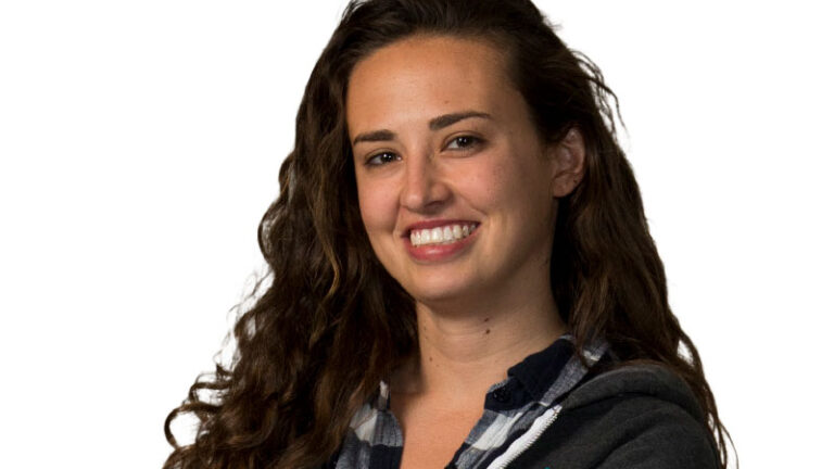 A woman smiling at the camera, wearing a plaid shirt and a dark jacket, against a white background.
