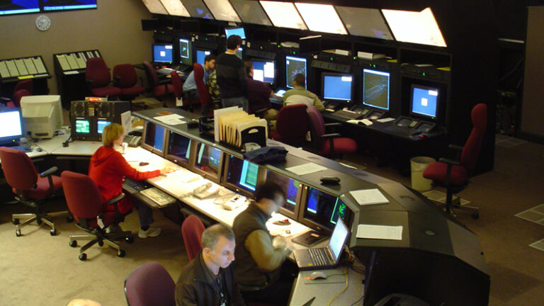 Control room with multiple operators at computer stations monitoring screens, with dim lighting and ergonomic office chairs.