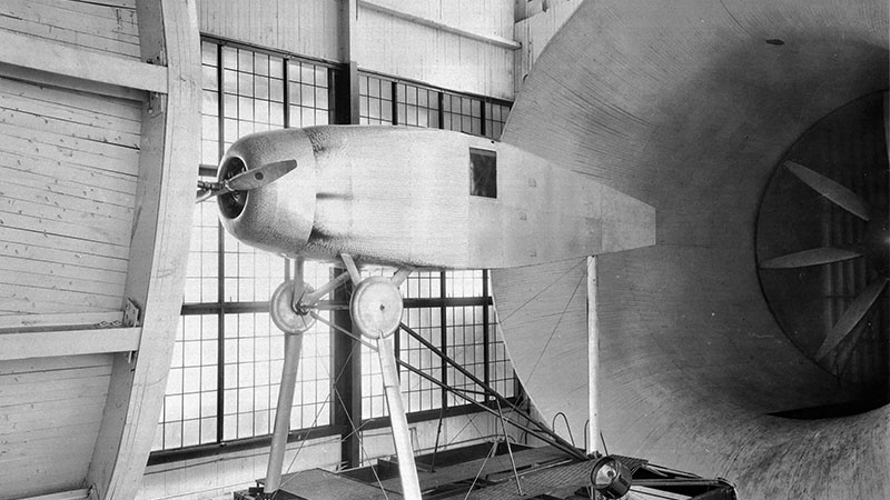 Vintage black-and-white photo of an early aerodynamic aircraft model displayed in front of a large wind tunnel.