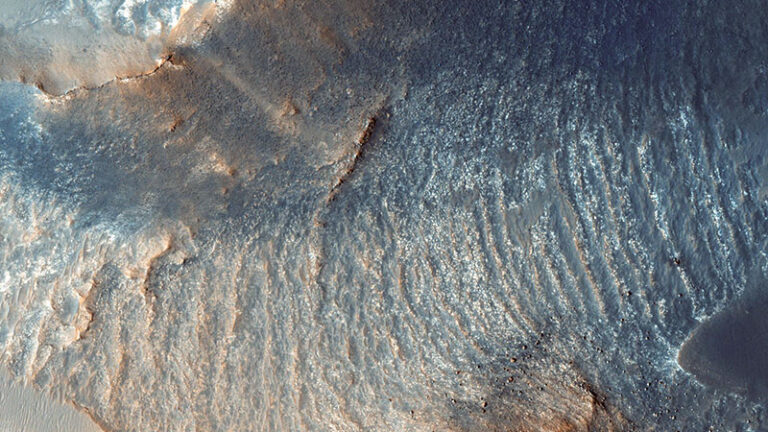 Aerial view of a rugged martian landscape featuring rippled textures and color variations ranging from deep blues to light browns.