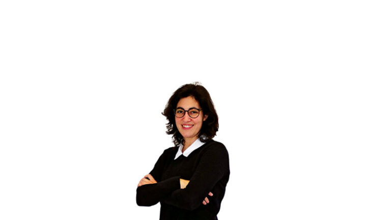 A woman with glasses wearing a black sweater over a white shirt, smiling with arms crossed, standing against a white background.