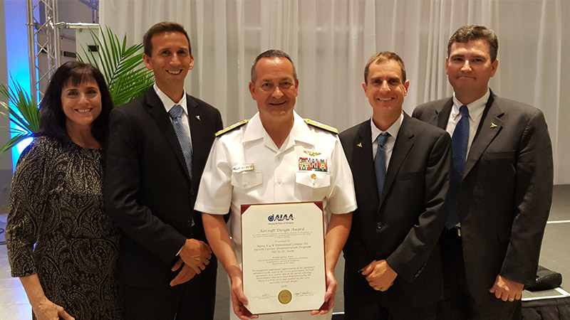 Five people, one in a naval uniform, standing with a framed certificate at a formal event with a backdrop of palm plants.