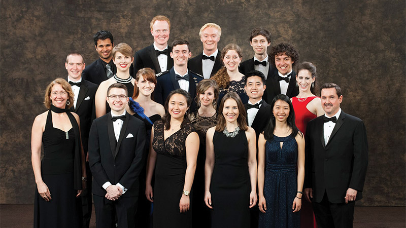 Group of fifteen people in formal attire posing for a photo against a dark backdrop.