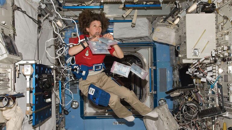 An astronaut floats in a space station, surrounded by equipment. They hold a food package while several other items float nearby.