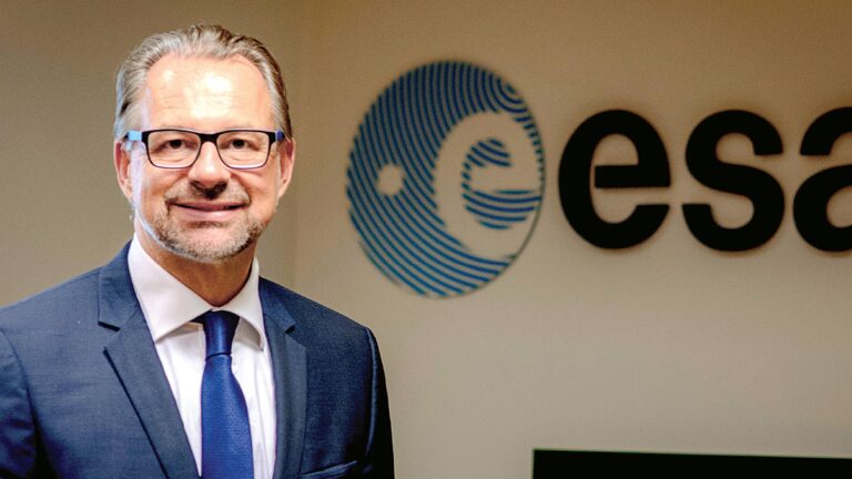 A man in a suit and tie stands in front of a wall displaying the European Space Agency (ESA) logo.