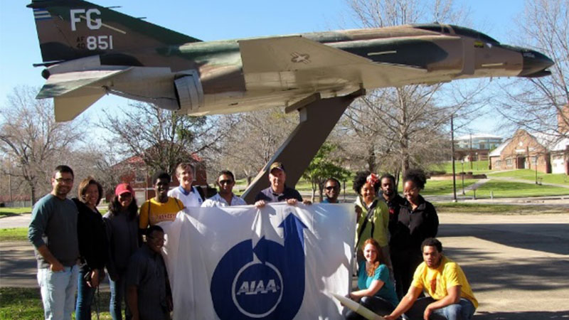 Group of students holding an aiaa banner in front of a mounted fighter jet on a sunny day on campus.