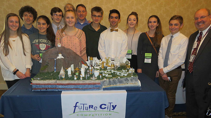 A group of students and two adults posing behind a model city at the future city competition.
