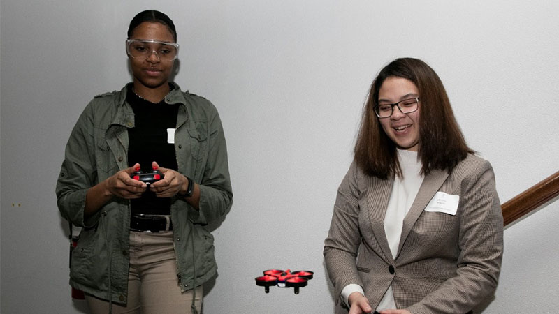 Two young women, one controlling a drone with a remote, smiling and engaging in a tech demonstration indoors.