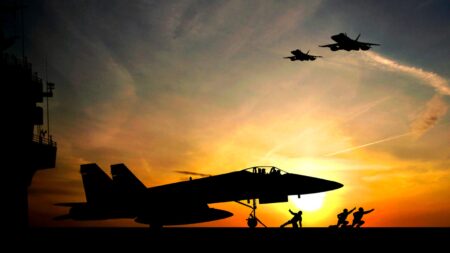Silhouette of a fighter jet on a runway against a sunset, with two jets flying overhead and people working near the aircraft.