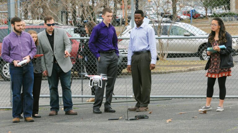 A group of five individuals, three men and two women, stand near a fence in a parking lot. Two men are operating drones while others observe. One woman holds a notebook and a tablet.