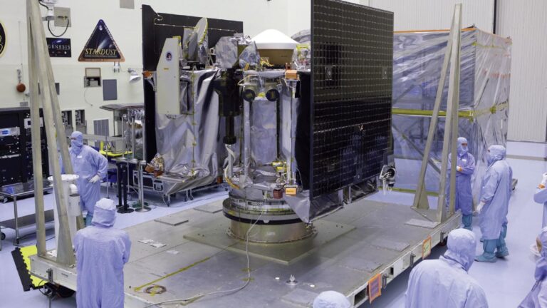 Technicians in cleanroom suits work on a large spacecraft with solar panels and equipment, positioned on a platform inside a laboratory.