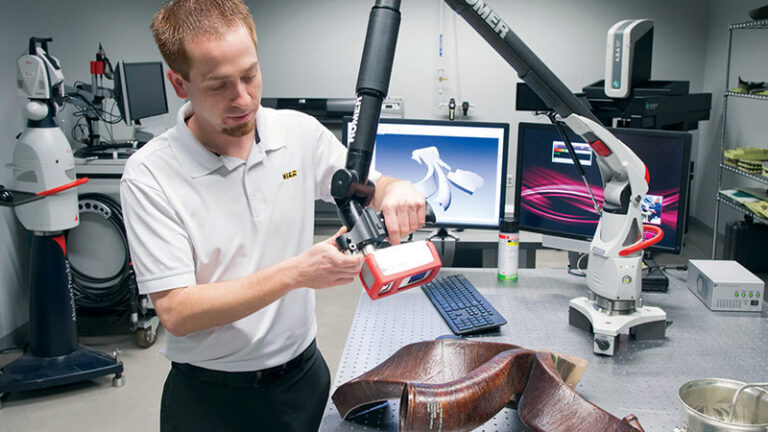 A technician uses a handheld 3d scanner on a metal part in a lab with monitors displaying scan data.