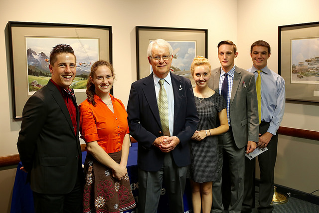 Five young adults and an elderly man posing together in a room with landscape paintings on the walls.