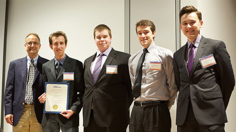 Five men, likely a team, posing with a certificate at a formal event; one is older and four are younger, all dressed in business attire.