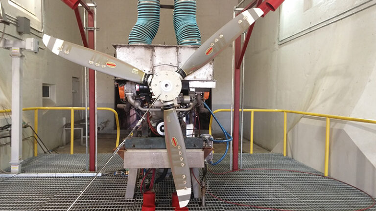 Aircraft engine on a test stand equipped with a multi-blade propeller and connected to various hoses, located inside an industrial testing facility.