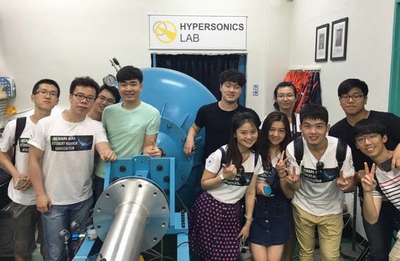 Group of nine students in a lab with a large blue machine, smiling and posing for the camera, some displaying peace signs. sign reads 