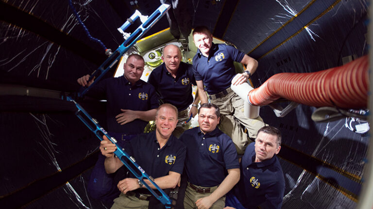 Six astronauts in blue nasa shirts pose inside the confined space of a spacecraft, smiling at the camera.