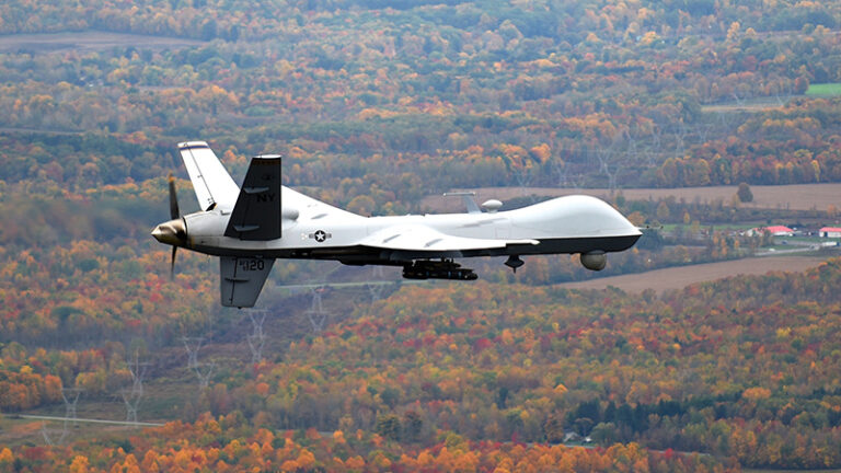 An mq-9 reaper drone flying over a colorful autumn landscape with scattered farmlands below.