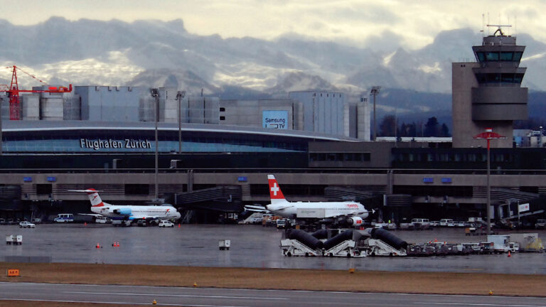 Zurich Airport with Swissair and Austrian Airlines planes parked at the terminal, set against a backdrop of snowy mountains and overcast sky.
