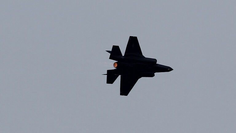 A silhouette of a fighter jet flying against a gray sky, with its afterburner ignited.