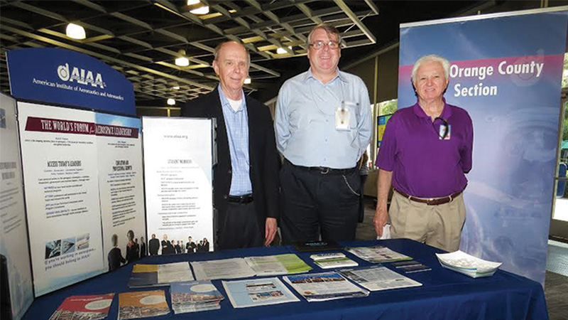 Three men standing by informational booths for the aiaa and orange county section at a conference.