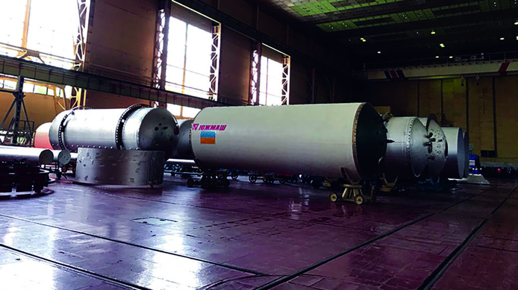 Large industrial warehouse storing multiple sections of rocket fuselages on transport dollies, with russian text visible on one segment.