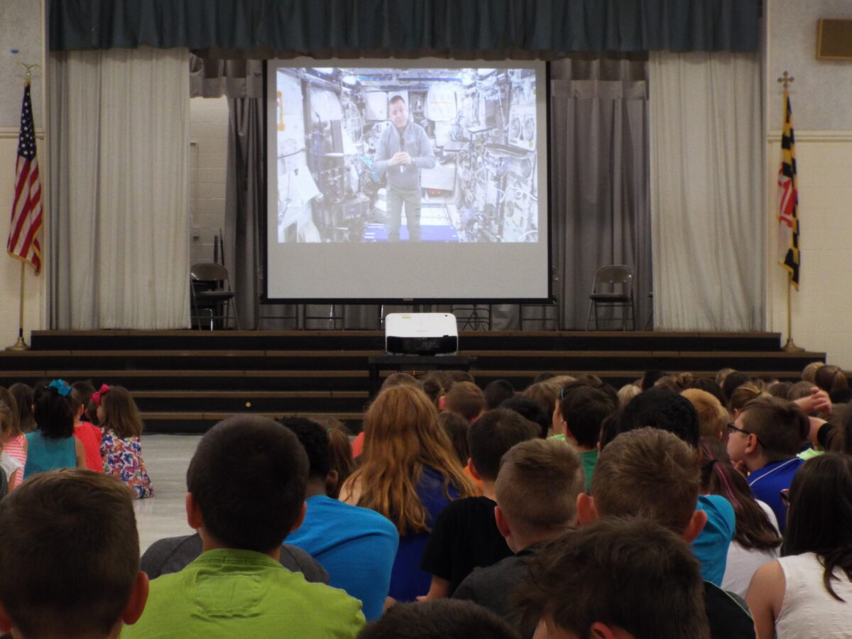Children watching an astronaut presentation on a projector screen in a school auditorium.