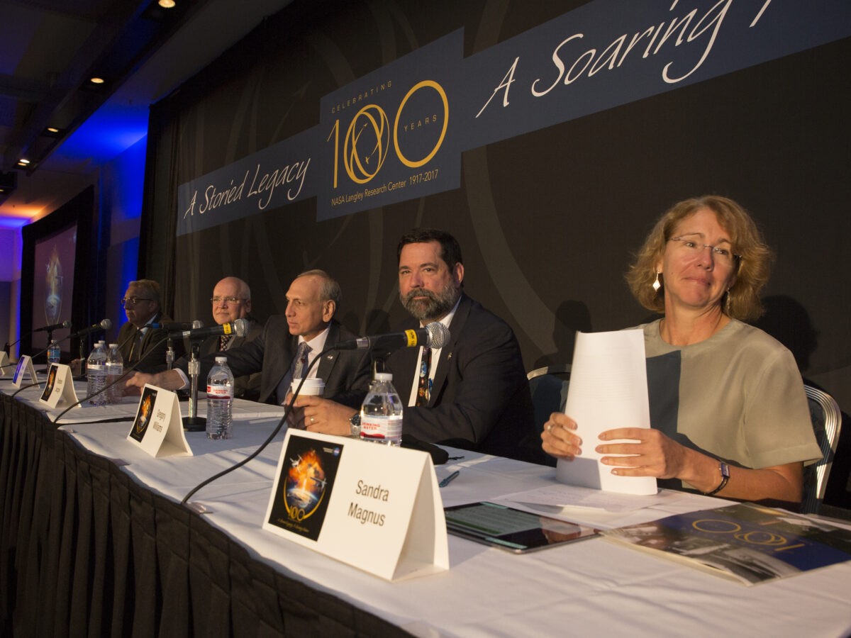 Panel of six diverse speakers sitting at a long table during a conference, each with a microphone and name tags displayed, in front of a banner reading 