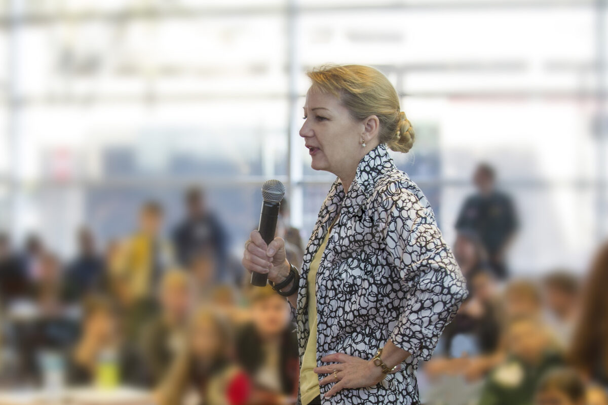 A woman speaking into a microphone in front of an audience, wearing a patterned shirt, with a blurred background.