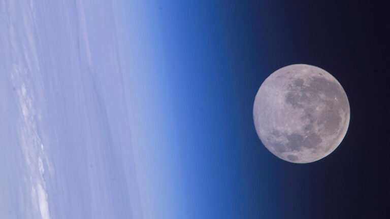 Full moon viewed against the backdrop of earth's atmosphere, showing a gradient from deep blue to black space.