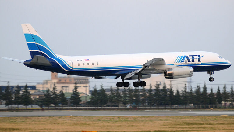 A blue and white at&t branded airplane landing, with its landing gear deployed, against a hazy sky and distant urban backdrop.