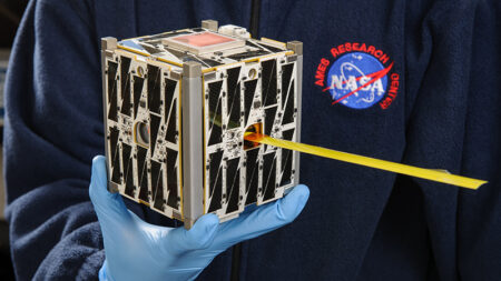 Person in a lab coat, wearing gloves, holding a small cubic satellite with solar panels, featuring a nasa research logo.