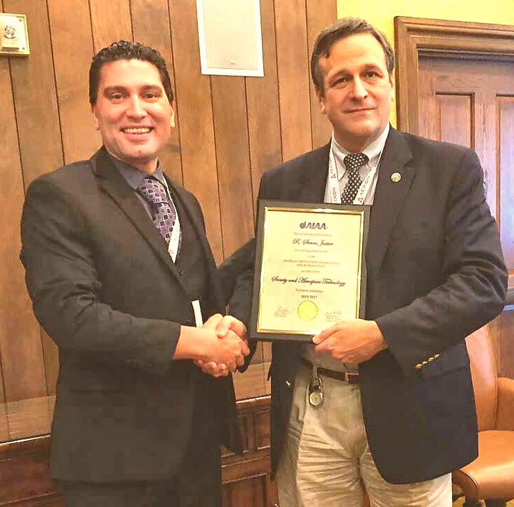 Two men shaking hands in an office, one holding a framed certificate, both smiling and dressed in business attire.