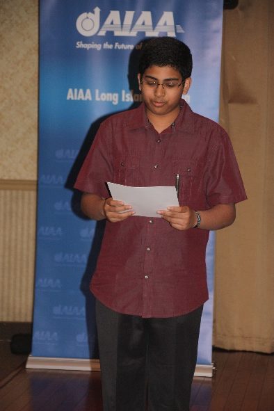 A young boy in glasses, wearing a red shirt, reads from a paper in front of an aiaa long island banner.