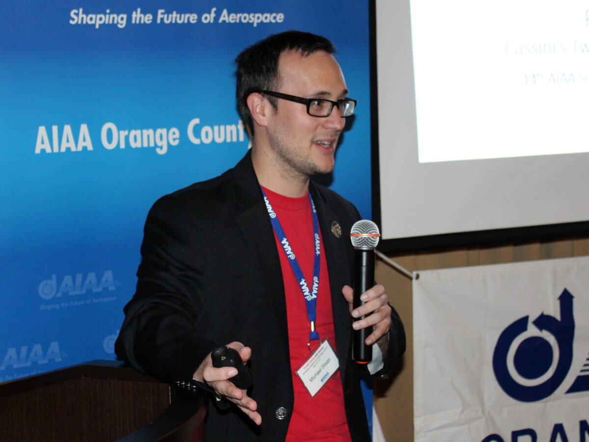 A man presenting with a microphone in front of aiaa orange county banners at a conference.