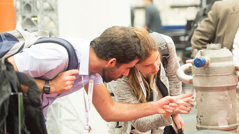 Two people examining a machine part at a trade show, with the man pointing at a detail and the woman looking intently.