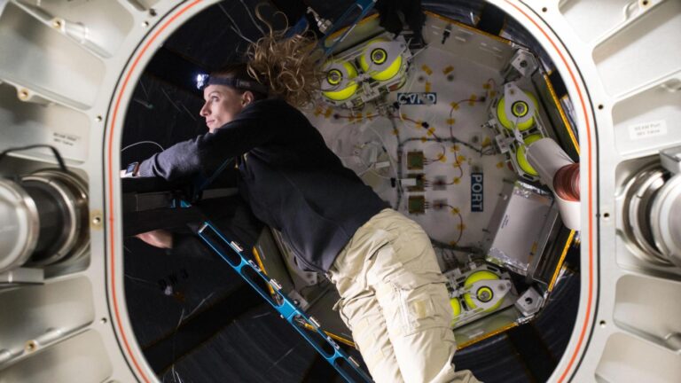 Person in a black shirt and beige pants floats in a space module, working with equipment inside a circular hatch. The interior features various instruments and storage units.
