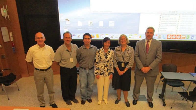 Six people smiling for a photo in a conference room with a large projection screen displaying a desktop background.