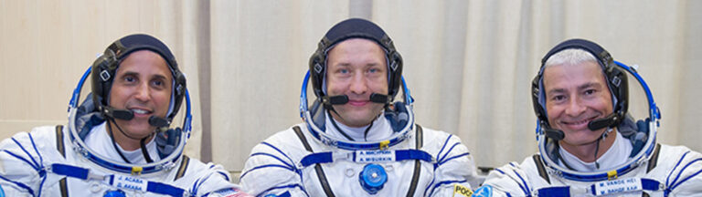 Three astronauts in spacesuits sitting side-by-side, smiling, with mission patches visible on their suits.