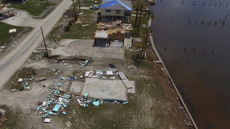 Aerial view of a coastal area showing damaged buildings and debris scattered around after a hurricane, with a flooded area nearby.